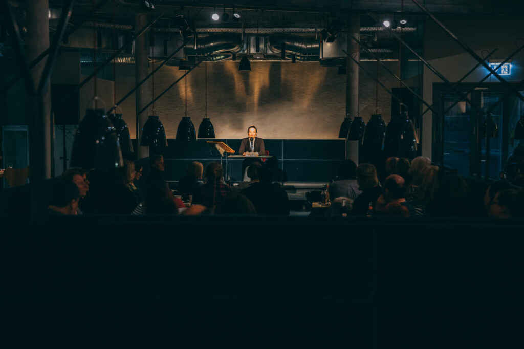 Auf dem Foto ist eine Bühne im Dunkeln auf der jemand in Anzug sitzt.