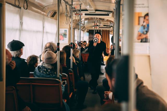 Auf dem Foto sitzen mehrere Menschen in einer Straßenbahn. Im Durchgang steht jemand und spricht in ein Megafon.