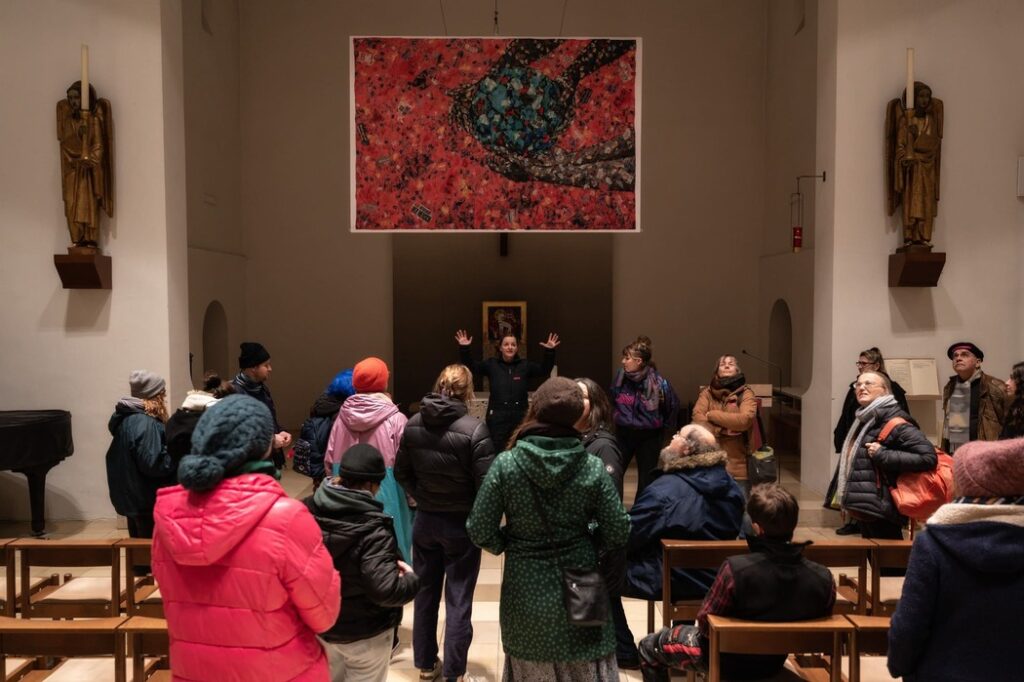 Auf dem Foto ist eine Gruppe versammelt in einer Kirche. Über ihnen hängt ein buntes Gemälde in roten Farben.