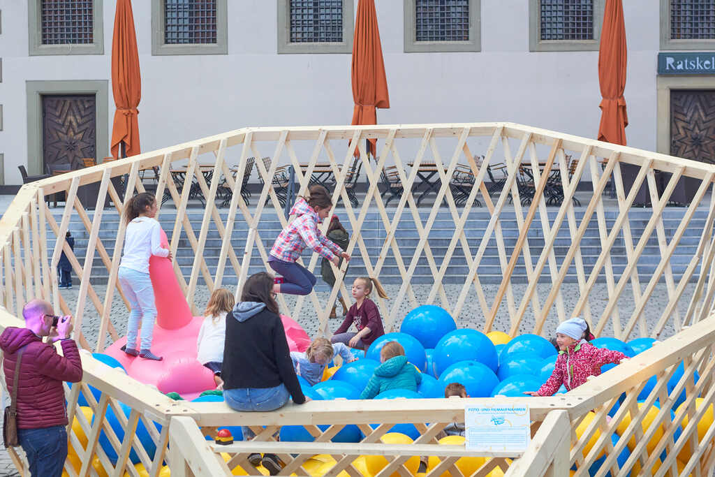 Auf dem Foto sind viele Kinder sitzend und hüpfend zwischen blauen und gelben Bällen sowie einem aufblasbaren Flamingo.