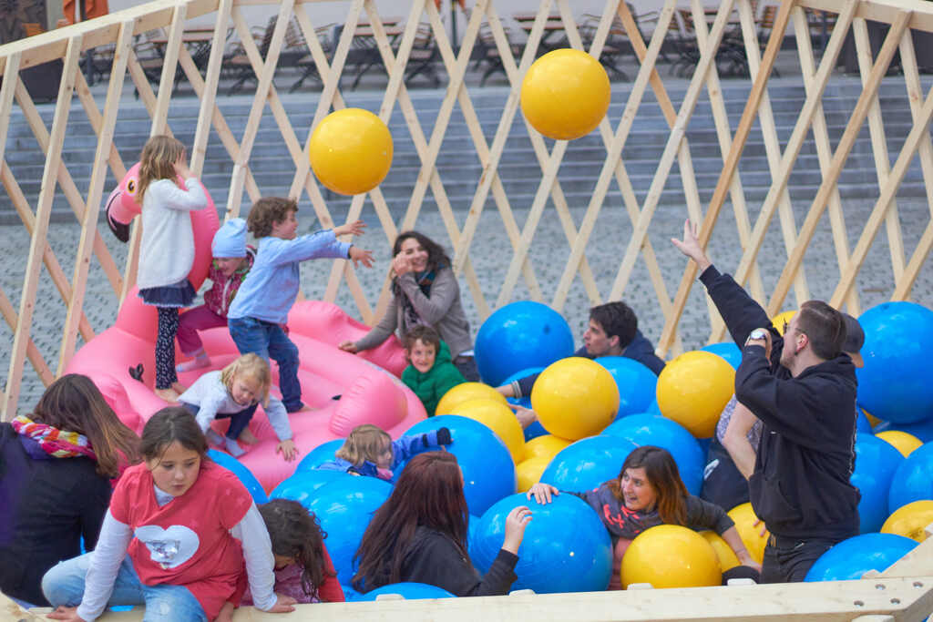 Auf dem Foto sind viele Kinder und Erwachsene zwischen blauen und gelben Bällen sowie einem aufblasbaren Flamingo. Sie werfen sich die Bälle zu und haben Spaß.