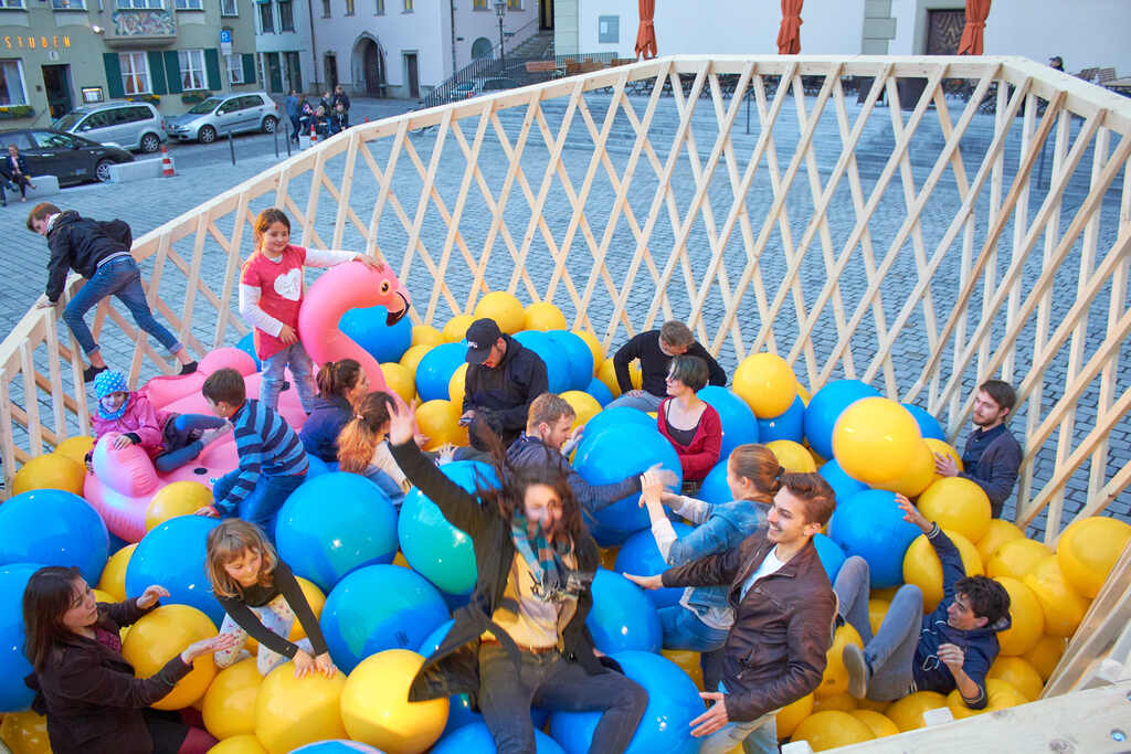 Auf dem Foto sind viele Kinder und Erwachsene zwischen blauen und gelben Bällen sowie einem aufblasbaren Flamingo. Sie toben umher und haben Spaß.