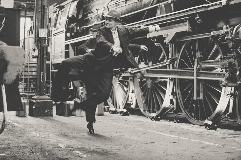 Auf dem schwarzweißen Foto ist eine Tänzerin in großem Anzug, die konzentriert in die Luft springt. Im Hintergrund steht ein alter Zug.