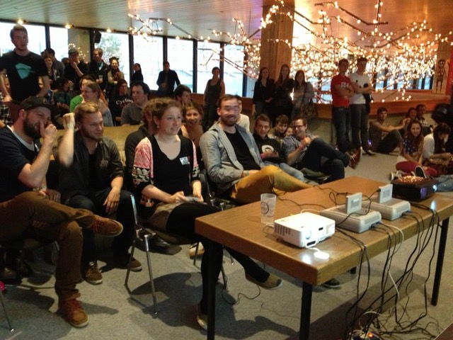 Auf dem Foto ist eine Gruppe an Menschen zu sehen, die hinter einem Tisch mit zwei Spielekonsolen und Beamern sitzen. Im Hintergrund sind viele Lichterketten zu sehen.