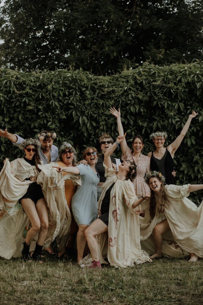 9 Personen aus dem Bluespots-Team stehen vor einer grünen Hecke auf dem Märchenzelt-Gelände und freuen sich üer ihren Erfolg. Alle strecken jubelnd die Arme von sich oder in die Höhe. Einige tragen Blumenkränze und weite Gewänder.