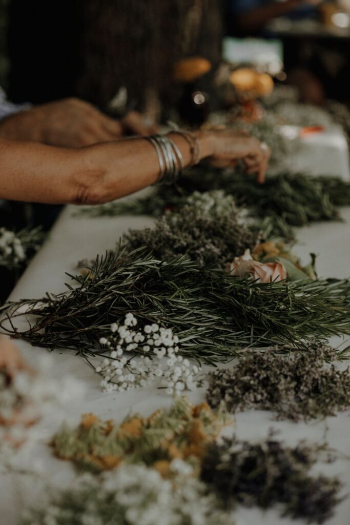 Zwei Personen, von denen nur die arme zu sehen sind, sitzen an einem Tisch und binden Blumenkränze. Der Tisch hat eine weiße Tischdecke. Auf ihm liegen die verschiedenen Pflanzen und Kräuter, die zum Blumenkranzbinden zur Verfügung stehen, zum Beispiel Schleierkraut und Rosmarin.