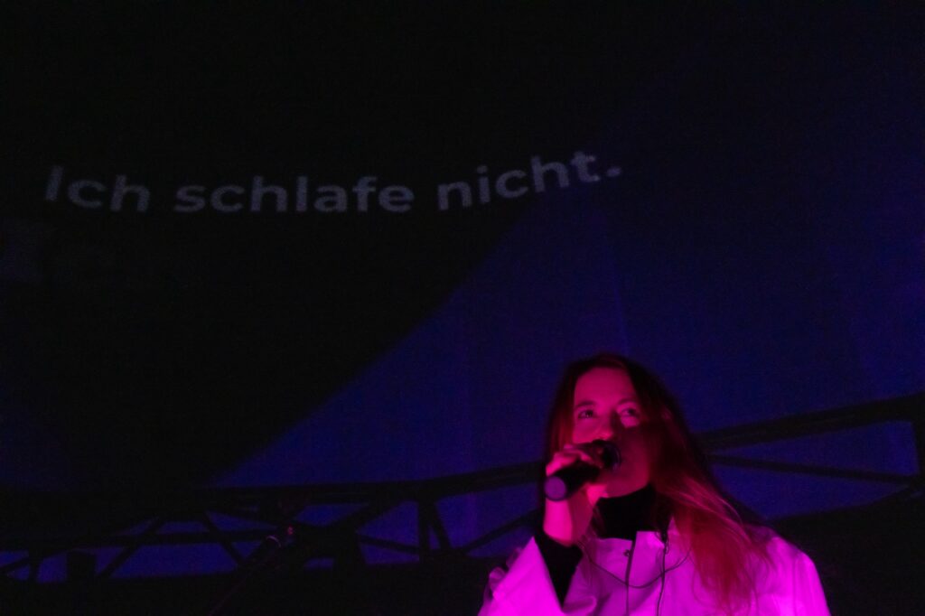 Die Sängerin Helene Schönfeld während der Performance. Hinter ihr wird ihr Text an die Wand projiziert. Dort steht: Ich schlafe nicht.