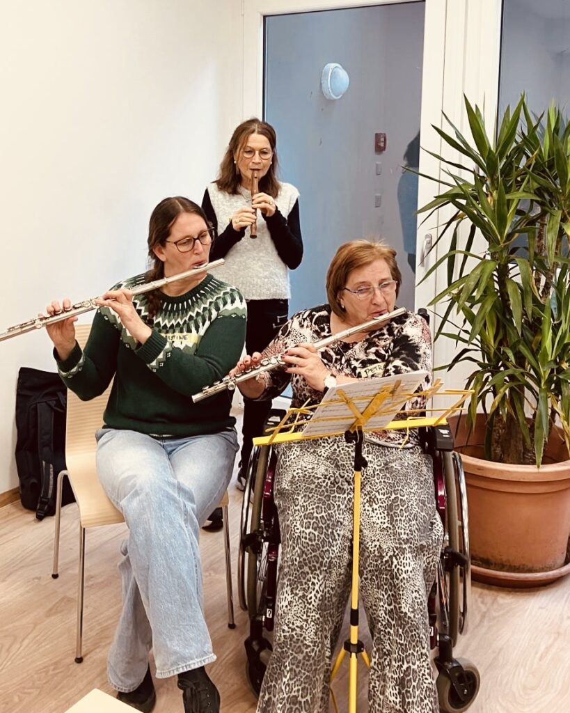 Drei Frauen musizieren in einem hellen Raum: zwei spielen Querflöte, eine Blockflöte. Eine der Flötistinnen sitzt im Rollstuhl, Noten stehen vor ihr.