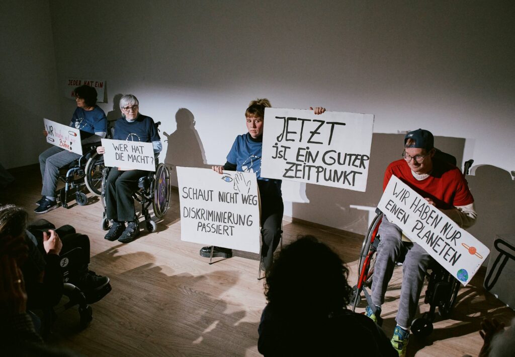 Die Schauspieler*innen Evi Schuster, Karin Lachenmair, Pia Altenried und Fabian Marquard halten während der Szene Zeitgeschehen Demo-Schilder in die Luft.