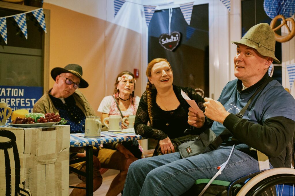 Die Schauspieler*innen Ralf Andersson Anja Neukamm, Stefan Kunz und Ina Wöhrle sitzen in der Szene Oktoberfest gemeinsam an einer Bierbank.